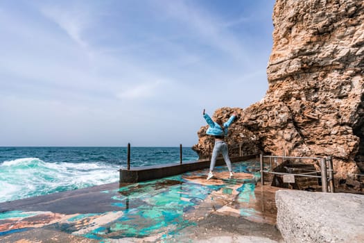 A woman in a blue jacket stands on a rock above a cliff above the sea and looks at the raging ocean. Girl traveler rests, thinks, dreams, enjoys nature. Peace and calm landscape, windy weather