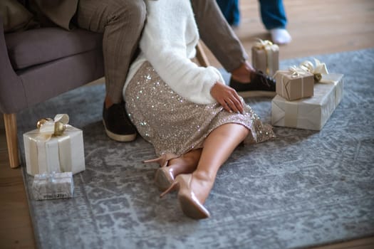 A romantic couple, a man sitting on the sofa and a woman on the carpet next to surrounded by gifts on a gray carpet. She is wearing a light skirt and beige high-heeled shoes