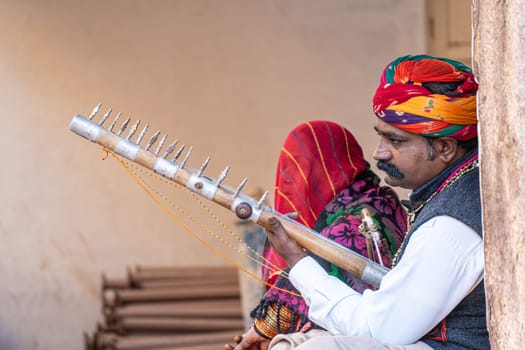 Jodhpur, Rajasthan, India - 26th Dec 2023: Rajasthani Indian musician sitting cross legged in colorful turban and white kurta pyjama local clothing in front of a brick wall at a mahal palace