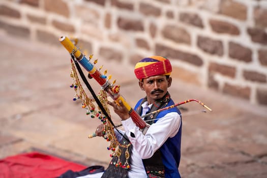 Jodhpur, Rajasthan, India - 26th Dec 2023: Rajasthani Indian musician sitting cross legged in colorful turban and white kurta pyjama local clothing in front of a brick wall at a mahal palace