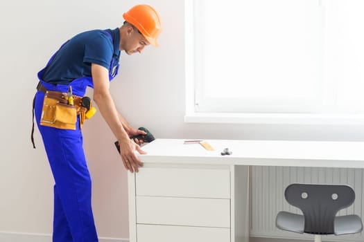 A man, a handyman in a blue shirt, collects a table, close-up. He assembles the table frame with a hex key while kneeling