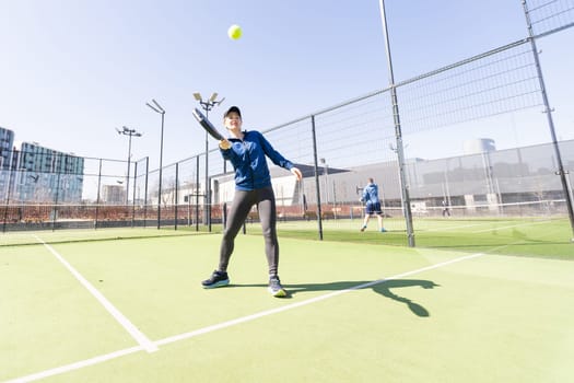 A girl in sportswear is training on a paddle tennis court. The girl is hitting the ball against the glass to make a rebound. Concept of women playing paddle. High quality photo