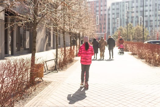 School-age sports girl is riding a scooter in the park on a walk on a sunny day. The child is warmly in hat. High quality photo