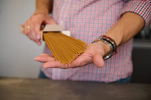 Close-up male hands holding a cardboard pack with integral spaghetti, standing at kitchen counter at home, preparing traditional Italian pasta for family dinner. Healthy food concept