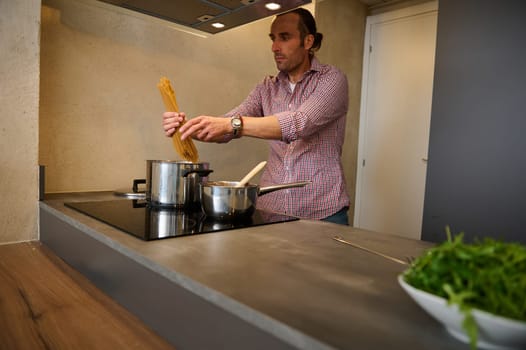 Young male chef cook preparing pasta at home kitchen, according to traditional Italian recipe, standing at electric induction stove, putting raw whole grain spaghetti into saucepan with boiling water.