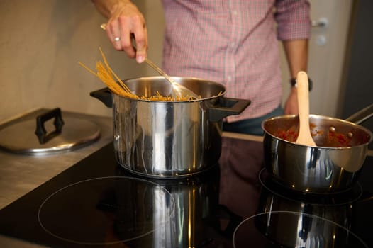 Man chef cook preparing Italian pasta at home. Cropped view of a young man standing at electric stove, putting raw whole grain spaghetti capellini into a stainless steel saucepan with boiling water.