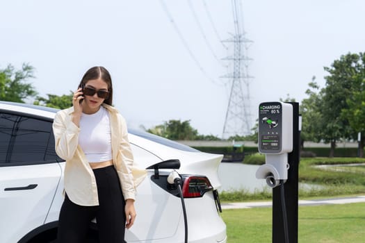 Young woman recharging EV car battery while talk on phone at charging station connected to electrical power grid tower facility as electrical industry for eco friendly vehicle utilization. Expedient