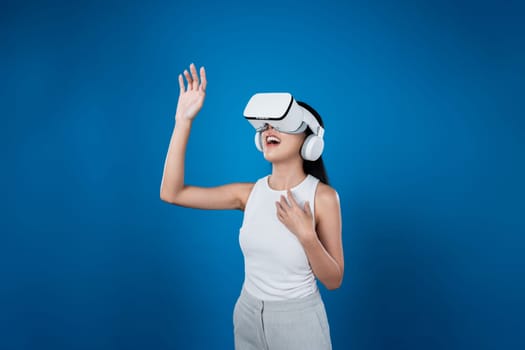 Smart female standing with blue background wearing VR headset connecting metaverse, futuristic cyberspace community technology. Elegant woman excited seeing generated virtual scenery. Hallucination.