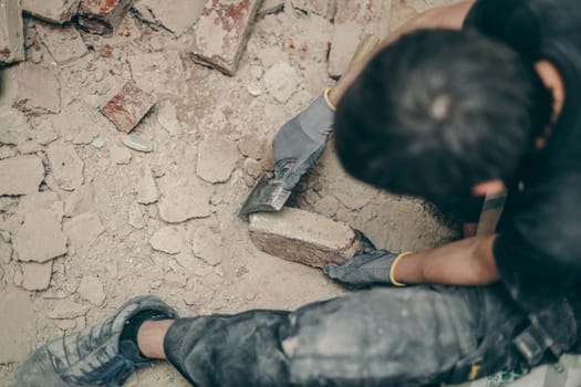 A young caucasian brunette man in black work clothes sits on a small chair on the right and cleans the bricks from dirt and old cement with an ax, top view close-up.Construction work concept.
