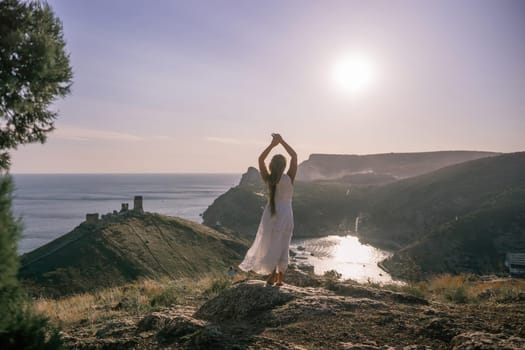 woman stands on a rocky hill overlooking a body of water. She is wearing a white dress and she is in a state of joy or celebration. Concept of freedom and happiness