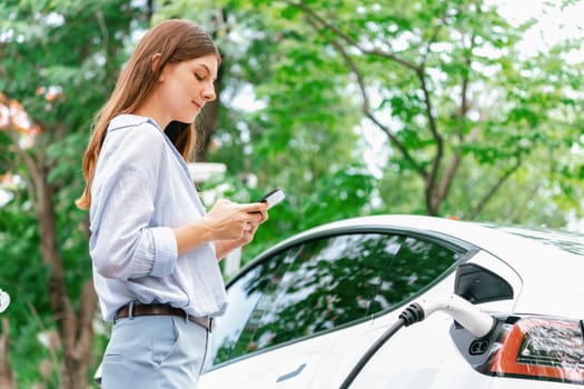 Young woman using smartphone online banking application to pay for electric car battery charging from EV charging station during vacation holiday road trip at national park or summer forest. Exalt