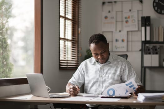 African American businessman working on paperwork, searching for information with laptop at home office.