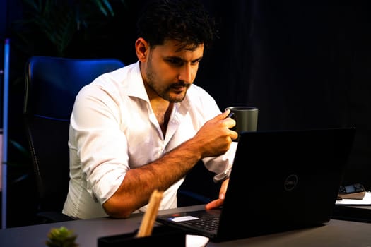 Working businessman with serious face holding coffee cup, researching information project on laptop at night time. Analyzing strategic business planning on database at neon light workplace. Sellable.