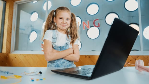 Girl using laptop programing software while looking at camera with arm folded with confident to celebrate successful project. Pretty child coding system while crossing arms in Stem class. Erudition.