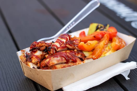 A paper container filled with a Californiastyle pizza topped with chicken, vegetables, and pizza cheese, ready to eat with a plastic fork as fast food