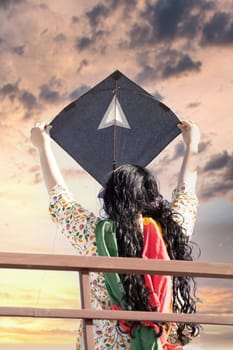 Young girl in traditional indian clothing holding black kite high above head launching it on sankranti republic independence day celebrations