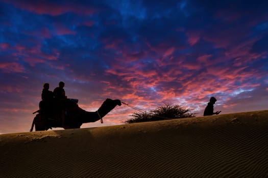 Silhouette of camel with two people sitting on it crossing over sand dunes in Sam Jaisalmer Rajasthan India