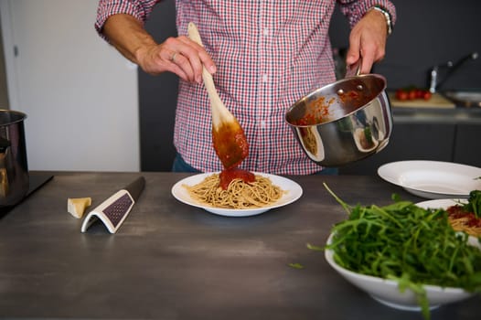 Cropped image of chef pouring tomato sauce on Italian pasta, plating up the dish before serving. Man cooking spaghetti for family dinner at home kitchen. Cuisine. Culinary. Epicure. Food concept