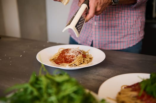 Close-up male hands grating cheese on freshly cooked Italian spaghetti with tomato sauce, preparing family dinner at home kitchen. Italian cuisine. Fresh ingredients on the kitchen table