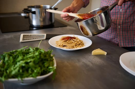 Young man cooking Italian pasta for dinner in the home kitchen. Close-up male hands holding a steel saucepan, pouring tomato passata sauce freshly cooked spaghetti,. seasoning the dish before serving