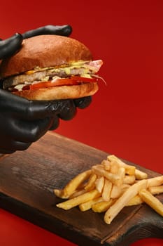 Hands in black rubber gloves holding tasty burger with chicken patty, fried bacon, fresh tomatoes, lettuce and cheese sauce, served with crispy fries on wooden board against red background