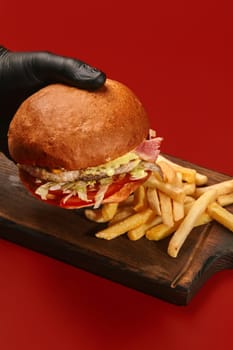 Hand in black gloves holding classic cheeseburger with juicy chicken patty, fried bacon, melted cheese and fresh vegetables served with side of golden fries on wooden board on red background