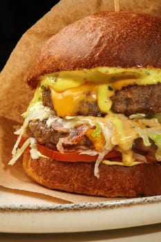 Close-up of appetizing burger in browned bun with two juicy beef patties, fried bacon slices, fresh tomatoes, lettuce and cheese sauce in craft paper wrapping on plate, against black background