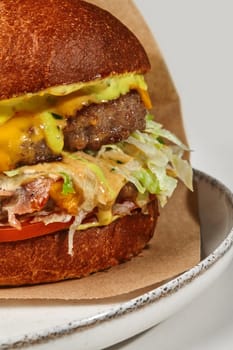 Close-up of delicious double hamburger in fluffy bun with two juicy beef patties, fried bacon slices, fresh tomatoes, lettuce and cheese sauce in craft paper wrapper on plate, against white background