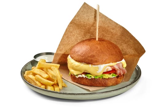 Appetizing chicken burger in fluffy browned bun with fried bacon slices, tomato, avocado and creamy cheese sauce wrapped in paper served with side of crispy fries on plate. Popular fast food concept