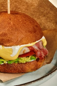 Close-up of appetizing chicken burger in fluffy browned bun with fried bacon slices, tomato, avocado and creamy cheese sauce wrapped in craft paper on plate. Popular fast food concept