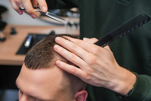 Hairstylist carefully cuts the brunette hair of the client with scissors at the barbershop.