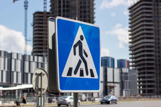 In front of a building under construction, there is a blue and white pedestrian crossing sign. The sky is visible in the background