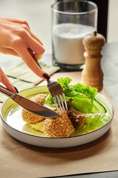 Female hands with perfect manicure using fork and knife to cut chicken patty in gold breading, served with side dish of mashed potatoes and green salad in cozy dining setting. Cropped shot