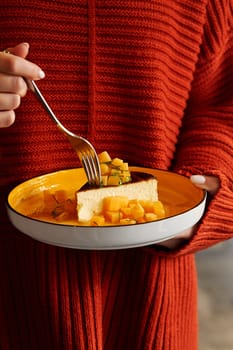 Woman in knitted red sweater holding plate with slice of creamy burnt basque cheesecake garnished with caramelized persimmon cubes and fresh mint, cropped shot. Moment of cozy tasting experience