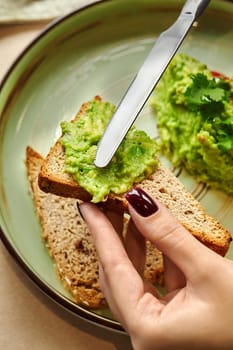 Female hands spreading vibrant piquant guacamole on slice of toasted whole grain bread. Tasty and healthy snack option