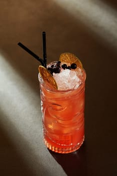 Glass of refreshing tropical Zombie cocktail with fruity and spicy notes on basis of rum with cinnamon syrup and grapefruit juice garnished with berries and dried lime slices on dark backdrop