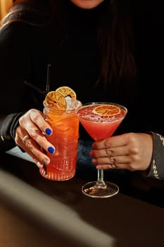 Two women clinking glasses with elegantly prepared chilled colorful cocktails with crushed ice garnished with dried citrus slices and berries, enjoying relaxed atmosphere in dimly lit bar