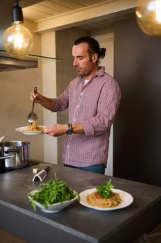 Caucasian handsome chef preparing Italian pasta at home kitchen, pouring tomato sauce over freshly boiled spaghetti. Fresh ingredients on countertop. Culinary. Epicure. Traditional Italian cuisine.