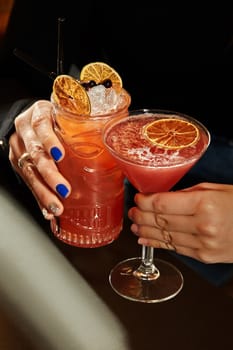 Close-up of female hands clinking glasses of beautifully garnished light cocktails with crushed ice, berries and dried citrus slices in dim bar