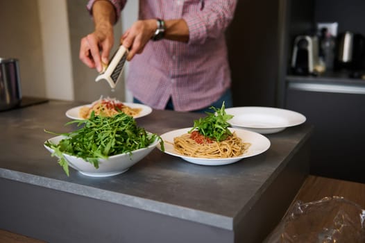 Close-up chef cooking dinner in stylish modern minimalist home kitchen interior, grating parmesan cheese over plate with freshly boiled spaghetti with tomato sauce. Ingredients on the kitchen counter