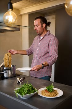 Young chef adding fresh green arugula leaves to Italian pasta with tomato sauce, garnishing the dish. A man preparing spaghetti for dinner in home kitchen. People and culinary concept. Italian cuisine