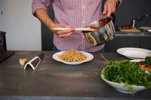 Close-up man pouring tomato sauce over plate with fresh spaghetti, standing at kitchen table with fresh organic ingredients. Cooking Italian pasta according to traditional Italian recipe