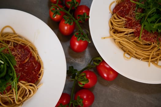 Flat lay white plates of spaghetti with tomato sauce and parmesan cheese, garnished with fresh green arugula leaves on dark background with a branch of cherry tomatoes. Classic Italian cuisine dish.