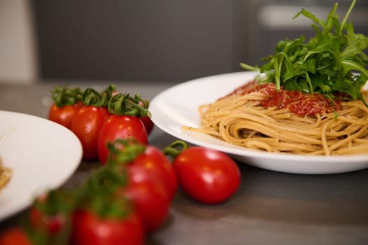 Food background. Italian spaghetti garnished with arugula leaves. Italian culinary, cuisine, culture and traditions. A bunch of red juicy ripe cherry tomatoes on the kitchen table on the foreground.