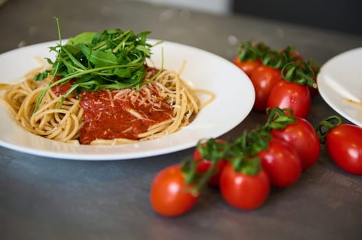 Food background. Still life with a white plate with Italian spaghetti capellini with tomato sauce, garnished with arugula green leaves and branch of red ripe organic tomatoes on the gray kitchen table