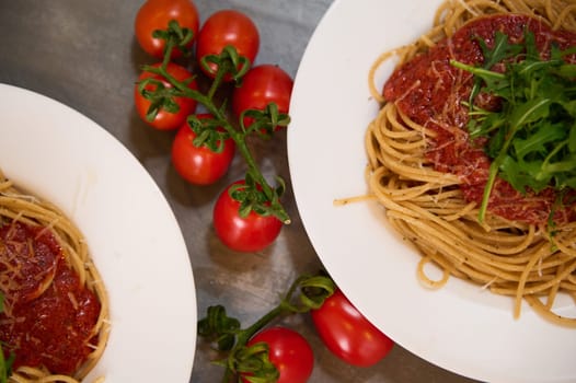 View from above. Partial view of two white plates with Italian spaghetti pasta with tomato sauce and arugula leaves, near a branch of fresh ripe organic cherry tomatoes. Food background