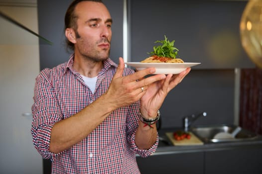 Caucasian handsome young adult man holding a white plate with Italian spaghetti capellini, standing in the home kitchen. Chef showing a served dish with Italian pasta. Italy. Food. Culture. Traditions