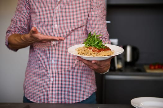 Close-up hands of young Italian chef showing a white plate with Italian spaghetti capellini, standing in the home kitchen. Chef showing served dish with Italian pasta. Italy. Food. Culture. Traditions