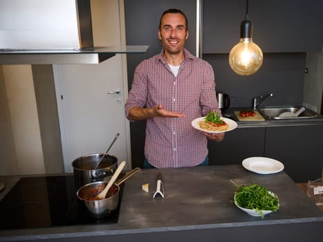 Attractive smiling young man 40s, showing at camera a plate with freshly cooked spaghetti pasta, standing at kitchen table with fresh ingredients and kitchen utensils. People. Italian food and culture
