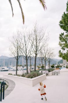 Little girl walks past flower beds with green trees on the embankment. Back view. High quality photo